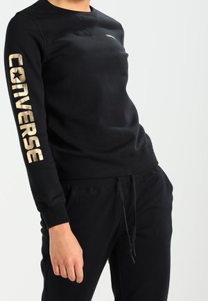 Svart sweatshirt med "CONVERSE" i stora beige bokstäver längs vänster ärm, bära med matchande svarta byxor med snörning.