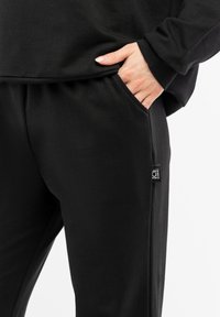 Black cotton sweatpants with side pockets and a small logo tag on the left side. Fabric is smooth, with a relaxed fit.