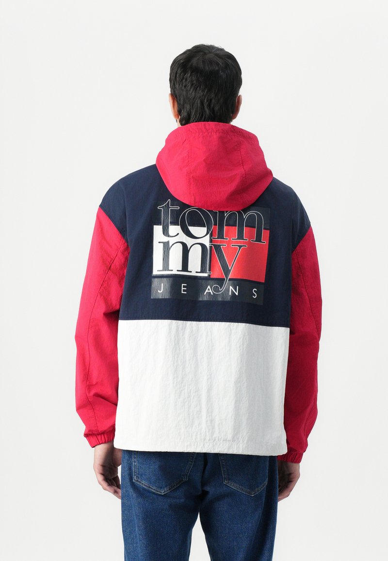 Farveblokket vindjakke med røde, marineblå og hvide sektioner; hætte, med "tommy JEANS" logoet tydeligt vist på ryggen.