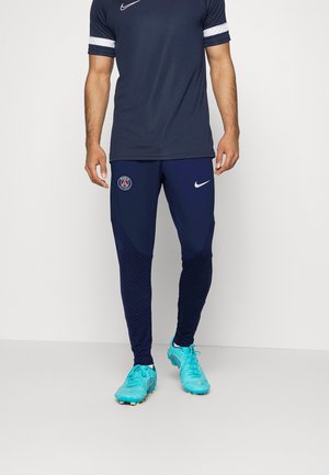 Mężczyzna ubrany w granatowy strój sportowy Paris Saint-Germain Nike z turkusowymi korkami, stojący na tle białego, jednolitego tła.