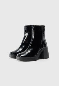 Bottines noires en patin brillant avec fermeture éclair, bout rond et talon épais. Texture lisse et finition brillante.