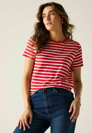 Joven mujer con cabello castaño ondulado, vestida con una camiseta de rayas rojas y blancas y vaqueros azules, mirando ligeramente de lado.