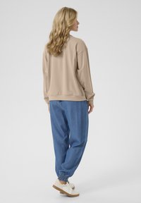 Felpa beige oversize con spalle scese, abbinata a joggers di denim blu larghi con polsini elastici. Sneakers bianche completano il look.