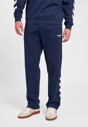 Mand iført marineblå hummel joggingbukser med hvide chevron-striber og beige sneakers står mod en ensfarvet baggrund.