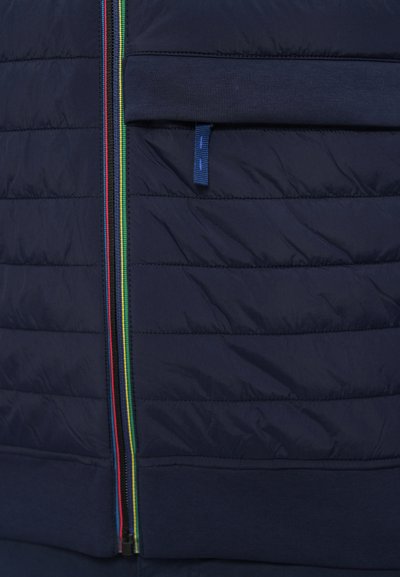 Tumesinine tikandiga jakk mitmevärvilise tõmbluku ja esitaskuga. Iseloomustab sile tekstuur ning kollase, punase ja rohelise kontrastsed aktsendid.