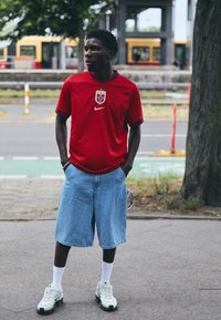 Maglietta rossa Nike con un emblema della Norvegia, pantaloni corti in denim blu e scarpe sportive bianche. Caratteristiche includono maniche corte e una vestibilità comoda.