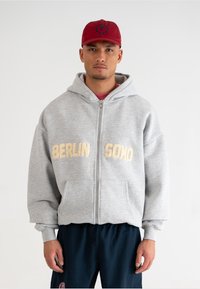 Felpa grigia con zip e la scritta "BERLIN SONO" ricamata in oro sulla parte anteriore, dotata di un ampio cappuccio e tasche a marsupio.