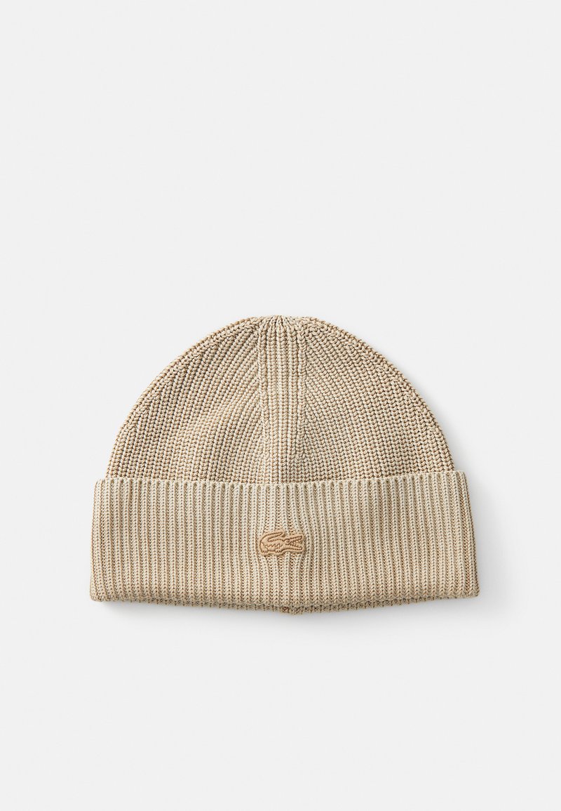 Bonnet beige tricoté avec une texture côtelée, comportant un logo Lacoste en cuir marron clair cousu sur le revers.