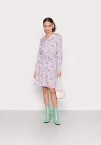 Vestido lilás com padrão floral, decote em V, mangas compridas, cintura ajustada, combinado com botas de tornozelo verde menta e uma pequena mala creme.