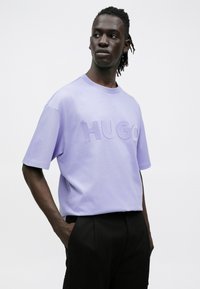 Νέος άνδρας με πλεξούδες στα μαλλιά που φοράει υπερμεγέθη λιλά μπλούζα με το γράμμα "HUGO" και μαύρο παντελόνι, στέκεται με τα χέρια στις τσέπες.