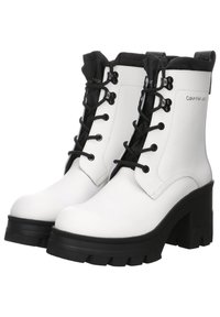 Bottes basses en cuir blanc avec talons et semelles épaisses noirs, lacets noirs, et logo Calvin Klein sur le côté.