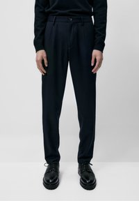 Marineblauwe, op maat gemaakte broek met een gladde textuur, voorzien van een voorplooi, riemlussen en een enkele knoopsluiting, gecombineerd met zwarte schoenen.