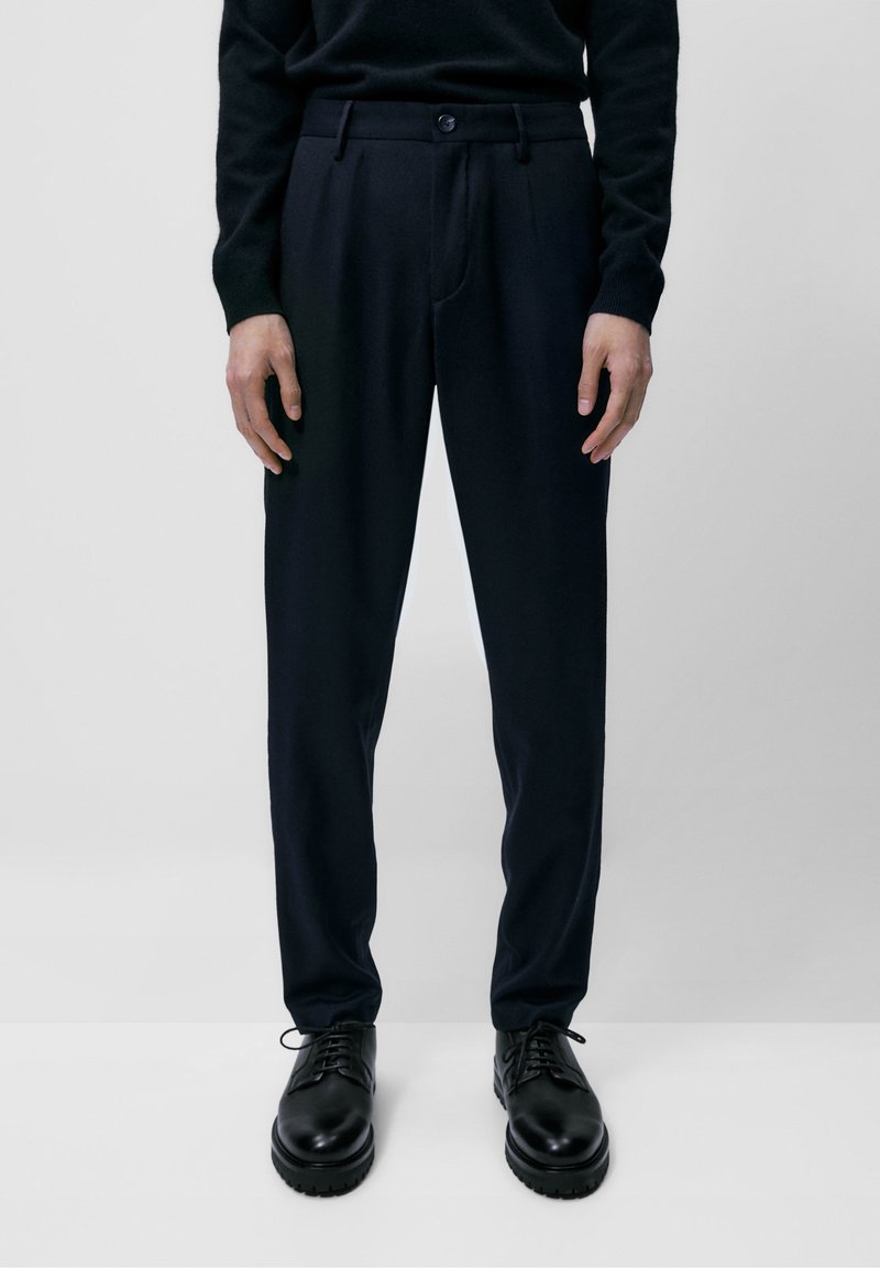 Marineblauwe, op maat gemaakte broek met een gladde textuur, voorzien van een voorplooi, riemlussen en een enkele knoopsluiting, gecombineerd met zwarte schoenen.