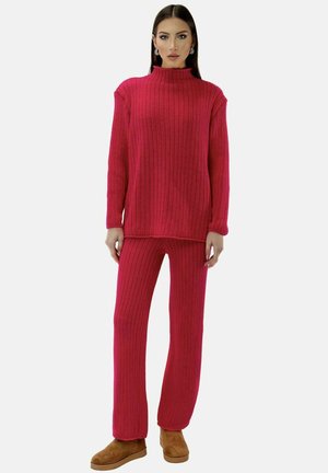 Kvinde iført en rød ribstrikket sweater og matchende bukser med brune ankelstøvler, stående mod en ensartet lys baggrund.
