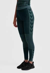 Mörkgröna atletiska leggings med en texturerad yta, med ett chevronmönster längs sidan och hög midja. Svarta sneakers.