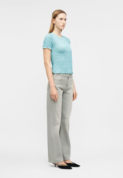 Γυναίκα όρθια, φοράει κοντομάνικο τοπ σε ανοιχτό μπλε με υφή, ανοιχτό γκρι τζιν με φαρδιά μπατζάκια και μαύρα χαμηλοτάκουνα παπούτσια, κοιτάζοντας αριστερά.