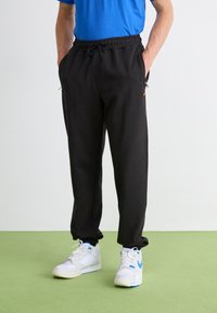 Pantalones de chándal negros con cinturilla elástica y cordón, bolsillos laterales, piernas ajustadas, combinados con zapatillas blancas con acentos azules.