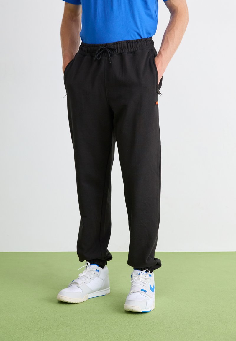 Pantalones de chándal negros con cinturilla elástica y cordón, bolsillos laterales, piernas ajustadas, combinados con zapatillas blancas con acentos azules.