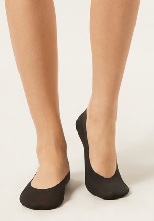 Nohy v černých ponožkách typu no-show na bílém pozadí, jedna noha položená celou plochou a druhá na špičkách.