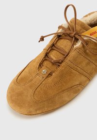 Sapato de camurça castanho com uma textura suave, apresentando um bico arredondado, fecho de atacador na frente e detalhes de costura contrastantes. Logotipo na lateral.