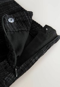 Svart zippered plagg med en texturerad ribbad krage, slät innerfoder och en knappdetalj nära dragkedjan.