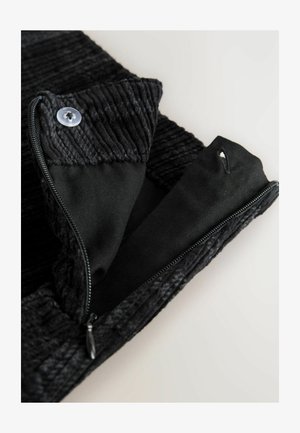 Svart zippered plagg med en texturerad ribbad krage, slät innerfoder och en knappdetalj nära dragkedjan.