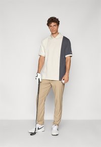Polo majica kratkih rukava u krem i tamnosivoj boji, s ovratnikom. Uparen s bež hlačama i bijelim Nike tenisicama. Drži golf palicu.