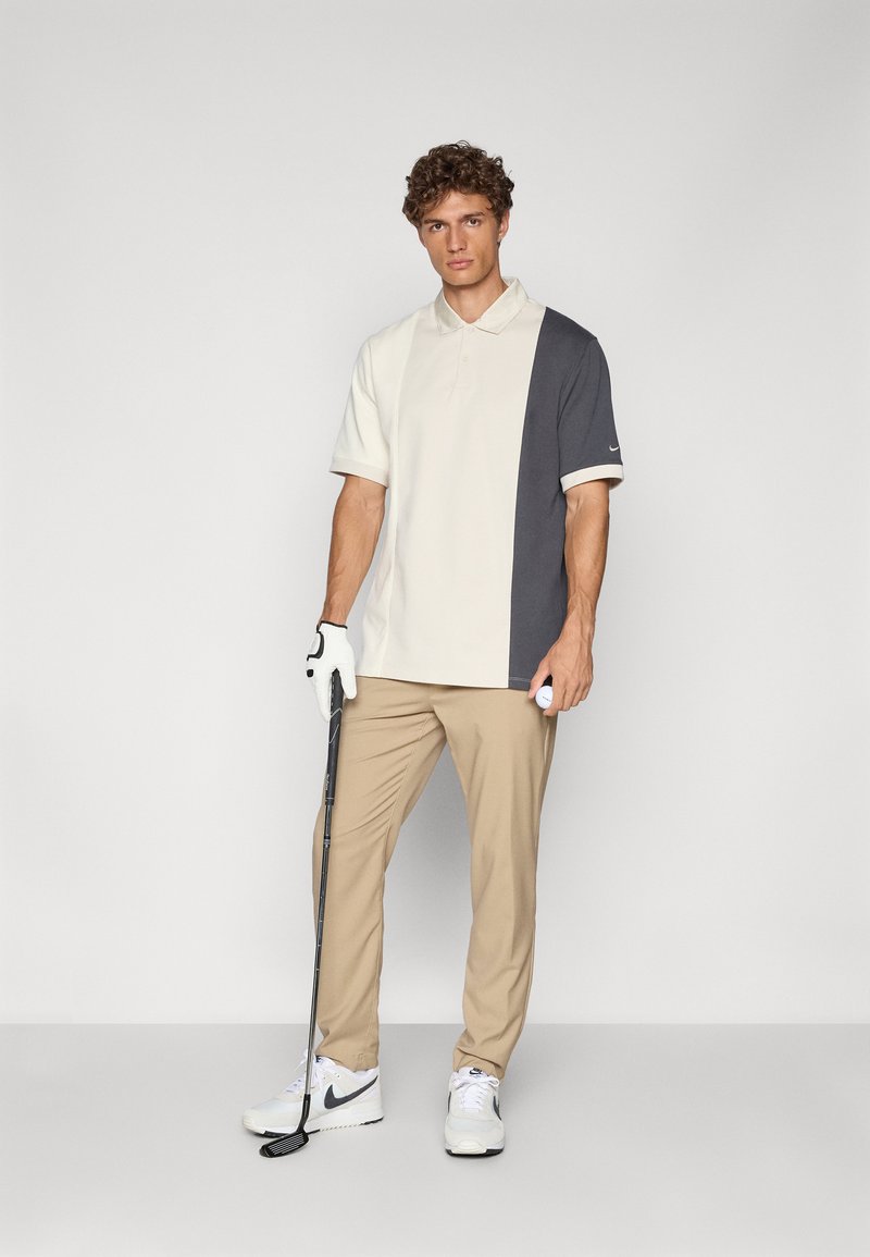 Polo de manga corta en color crema y gris oscuro, con cuello. Combinado con pantalones beiges y zapatillas Nike blancas. Sosteniendo un palo de golf.