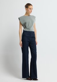 Světle šedý top s unikátním draped designem a zdobeným výstřihem, kombinovaný s tmavě modrými rozšířenými džíny, které mají zadní kapsy a viditelné švy.