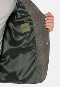 Eine Person trägt ein kariertes Blazerjackett und hält das Jacket offen, um das dunkelgrüne Satinfutter mit dem Text "BENVENUTO" und "COLLEZIONE #2" zu zeigen.