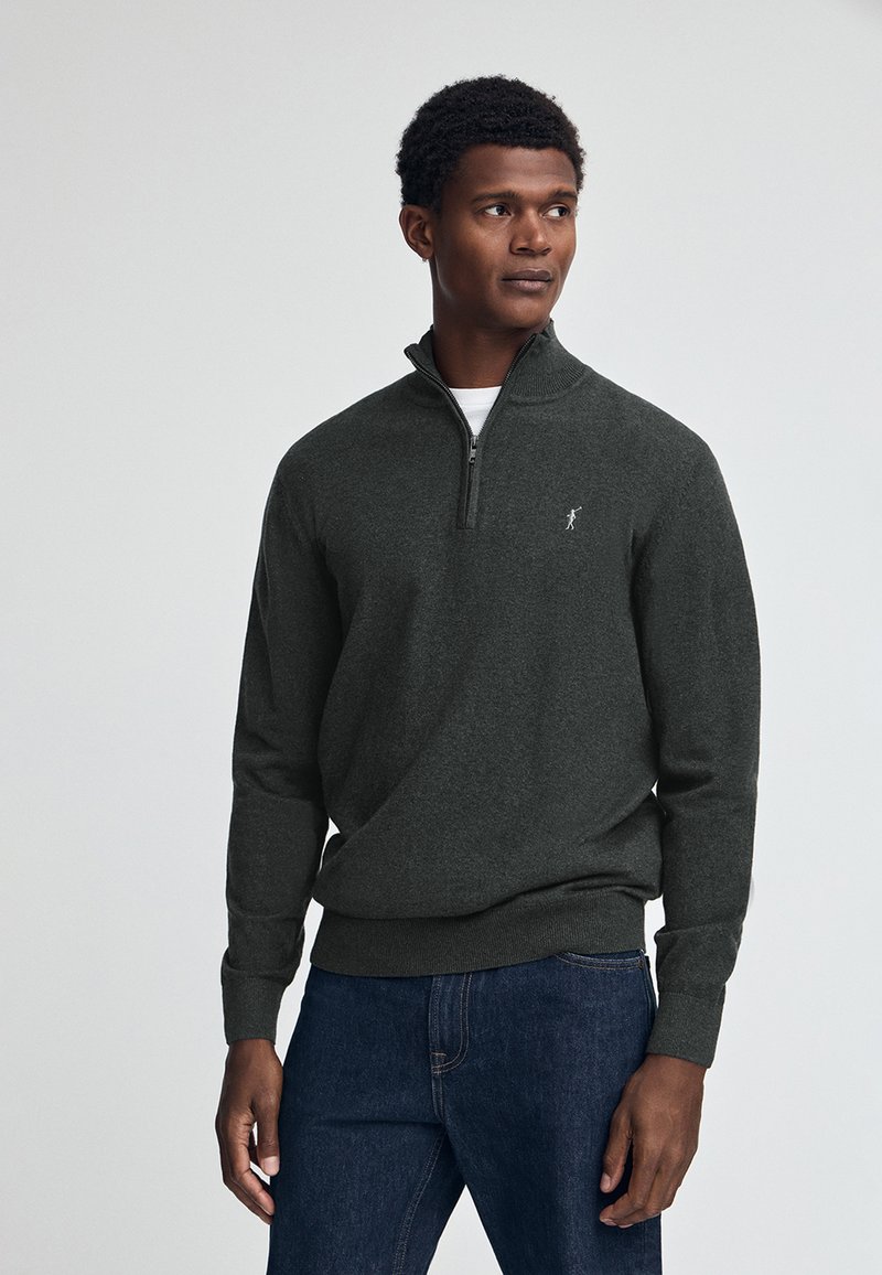 Mörkgrå zip-up-tröja med ribbade ärmslut och nederkant, med en liten logo på bröstet. Bärs över en vit skjorta med jeans.