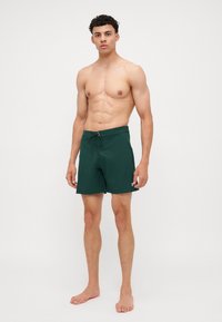 Grønne svømmeshorts med snøring i livet, laget av lett materiale, har en liten logo som accent på venstre lår.