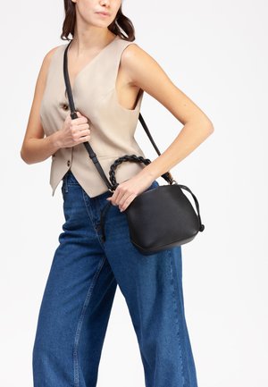 Donna che indossa un gilet beige senza maniche e jeans blu, tiene in mano una borsa nera con superficie strutturata, manico intrecciato e tracolla.