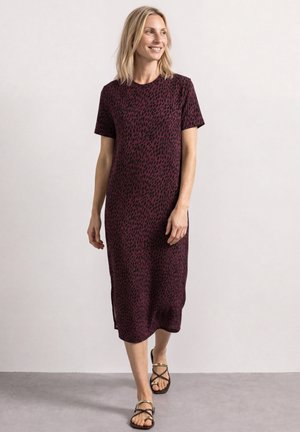 Frau mit schulterlangem blondem Haar, die ein lila-schwarz gemustertes Midikleid und schwarze Sandalen trägt, geht auf einem grauen Boden.