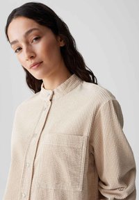 Beige corduroy shirt met een hoge kraag, knopen aan de voorkant en een borstzak. Heeft een gestructureerd, ribbelpatroon en een relaxte pasvorm.