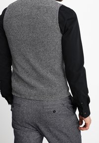 Chaleco texturizado gris sobre una camisa negra de manga larga, combinado con pantalones gris oscuro que tienen bolsillos traseros y detalles con botones.