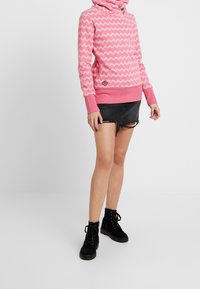 Felpa rosa con motivo zigzag bianco, tasche a marsupio e colletto alto; abbinata a pantaloni corti neri strappati e stivali neri stringati.