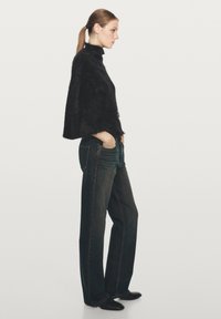 Kvinna i svart pälsmantel, mörka vida jeans och svarta spetsiga skor, som står i profil mot en enfärgad ljus bakgrund.