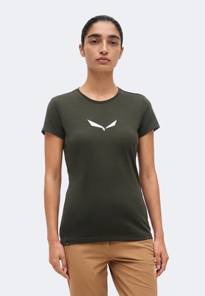 Joven con cabello oscuro recogido, vistiendo una camiseta ajustada verde oscuro con un logo blanco de un pájaro y pantalones color beige, de pie sobre un fondo sencillo.