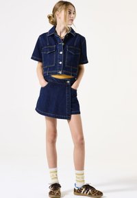 Denim cropped jack met korte mouwen en twee borstzakken, gecombineerd met een denim wikkelrok. Beide zijn donkerblauw met contrasterende stiksels.