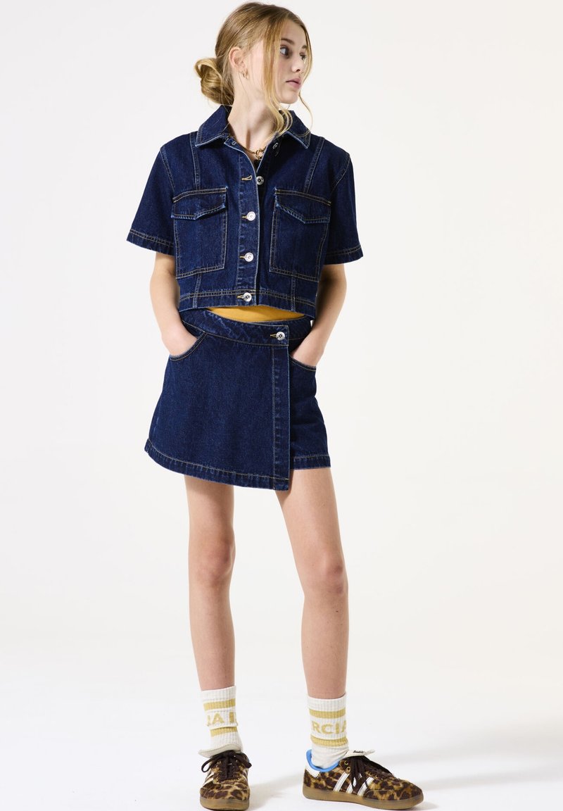 Denim cropped jack met korte mouwen en twee borstzakken, gecombineerd met een denim wikkelrok. Beide zijn donkerblauw met contrasterende stiksels.