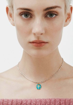 Collar de cadena de plata con un colgante de turquesa con diseño de corazón, descansando sobre piel desnuda, combinado con una blusa texturizada rosa.