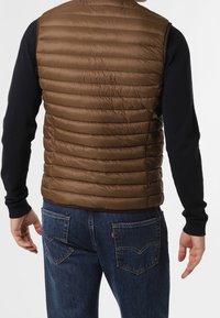 Brauner Steppwesten mit ge quilteter Textur, ärmelloses Design, getragen über einem schwarzen Langarmshirt, kombiniert mit dunklen Jeans.