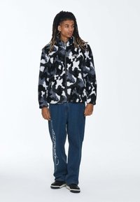 Kurtka polarowa w czarnym, białym i szarym kamuflażu z wysokim kołnierzem i zamek błyskawicznym. W połączeniu z luźnymi niebieskimi dżinsami.