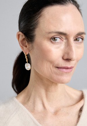 Les boucles d'oreilles pendantes dorées présentent une chaîne texturée avec un grand pendentif en perle de forme irrégulière. L'arrière-plan est neutre.
