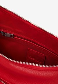 Interno di una borsa rossa in pelle con zip argentata, dotato di una piccola tasca con zip e un'etichetta. Superficie testurizzata con una finitura liscia.