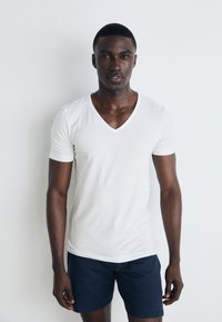 T-shirt blanc à manches courtes et décolleté en V présenté sur un modèle, assorti à un short bleu marine, le tout sur fond clair uni. Texture de tissu lisse.