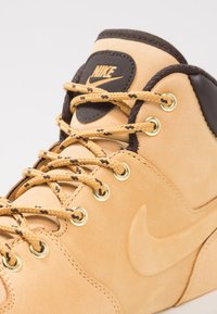 Baskets montantes en cuir tan avec des accents noirs, œillets dorés et lacets tressés. Présente un logo Nike en relief sur la languette et sur le côté.