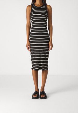Kvinde iført en ærmeløs, sort og hvid, vandret striped knælængde kjole med sorte slide sandaler, stående mod en ensfarvet baggrund.