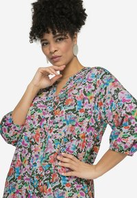 Robe fleurie en tissu léger avec une base rose, présentant des couleurs vives, un décolleté en V et des manches ballons. Comprend des poches avant.
